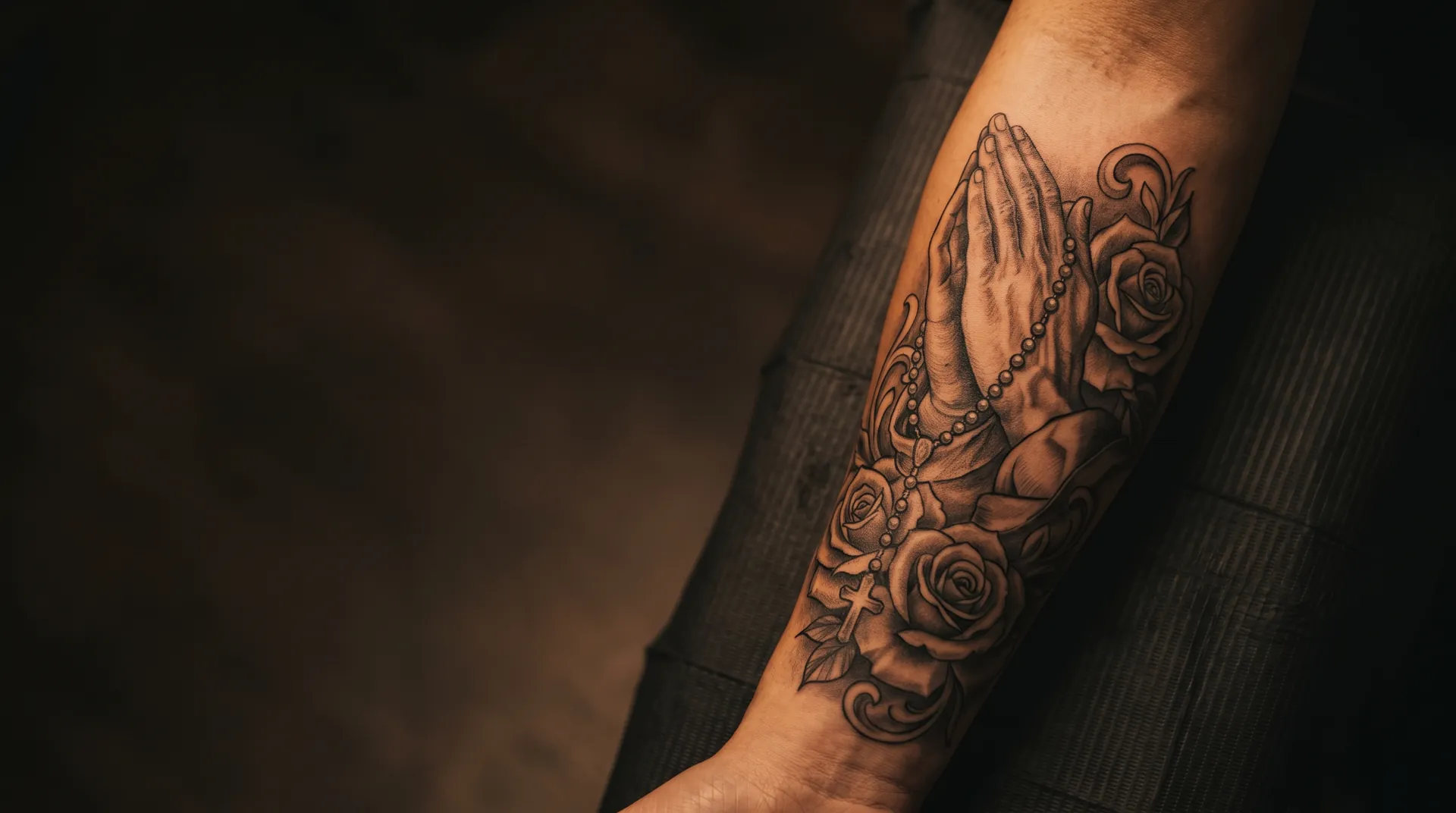 Chicano black and grey tattoo of praying hands with a rosary and roses on a forearm